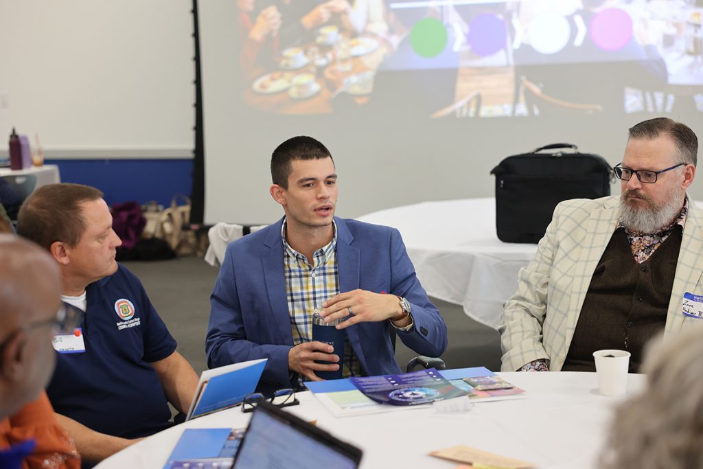 Roach sitting around a table with community partners sharing ideas. He is wearing a plaid shirt and blue blazer. 