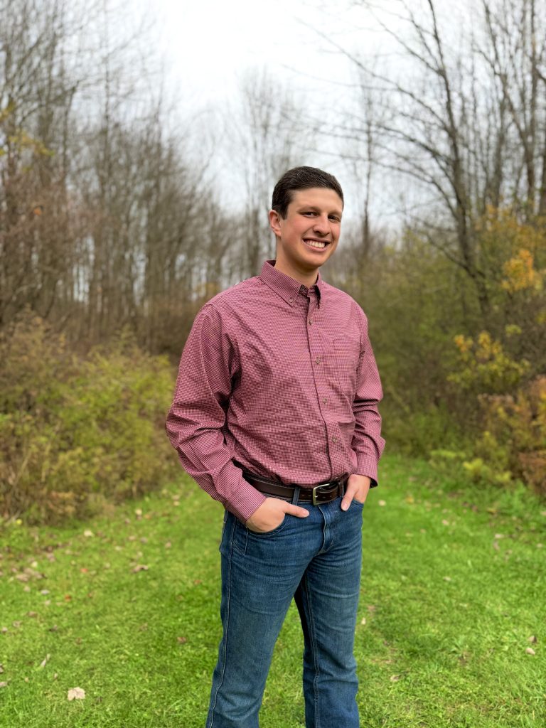 John Pertell standing outdoors in a scenic landscape, wearing a salmon colored dress shirt and dark blue jeans.