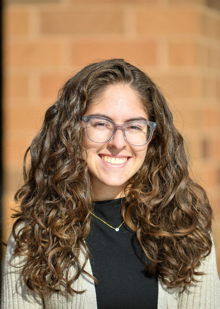 Carly Pimpinella headshot. She is outside, wearing beige sweater, black shirt, hair is curly and she is wearing clear glasses.