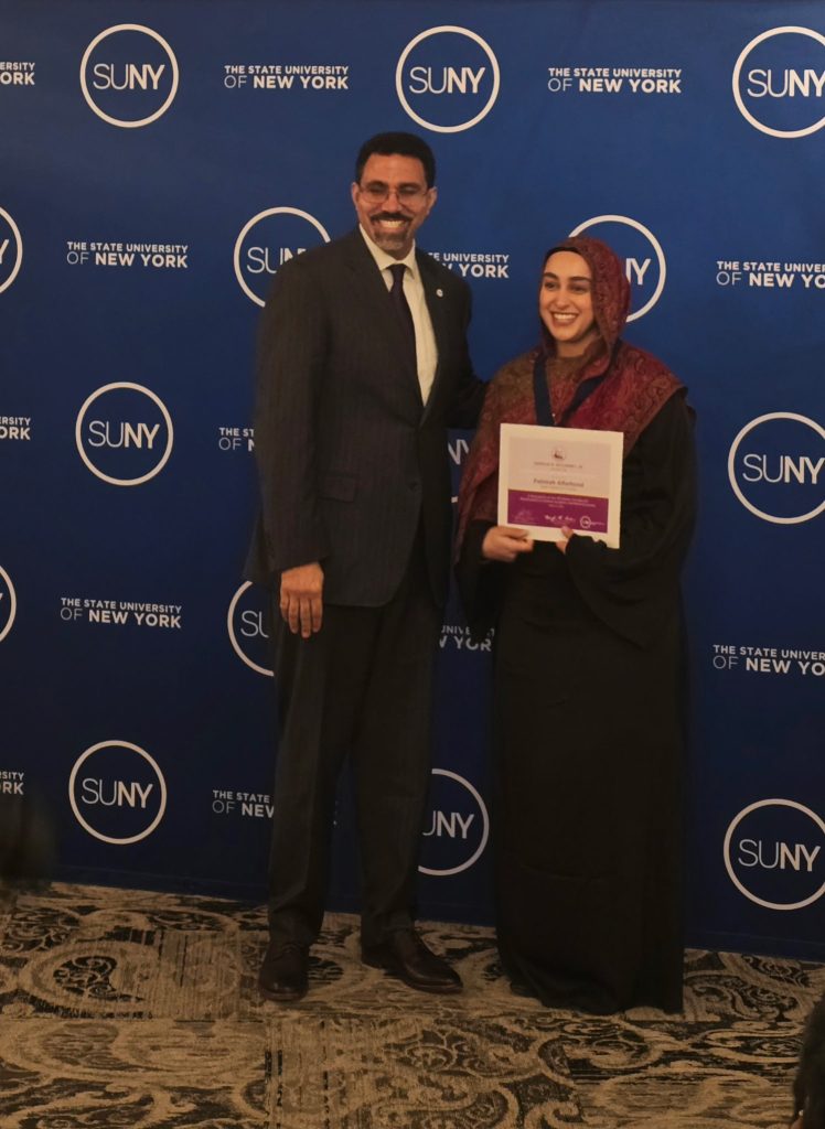SUNY Chancellor John B. King Jr in Black suit with black tie, wearing glasses. Fatimah is next to him in black dress and red patterned hijab holding her award. Behind them is a SUNY system backdrop.