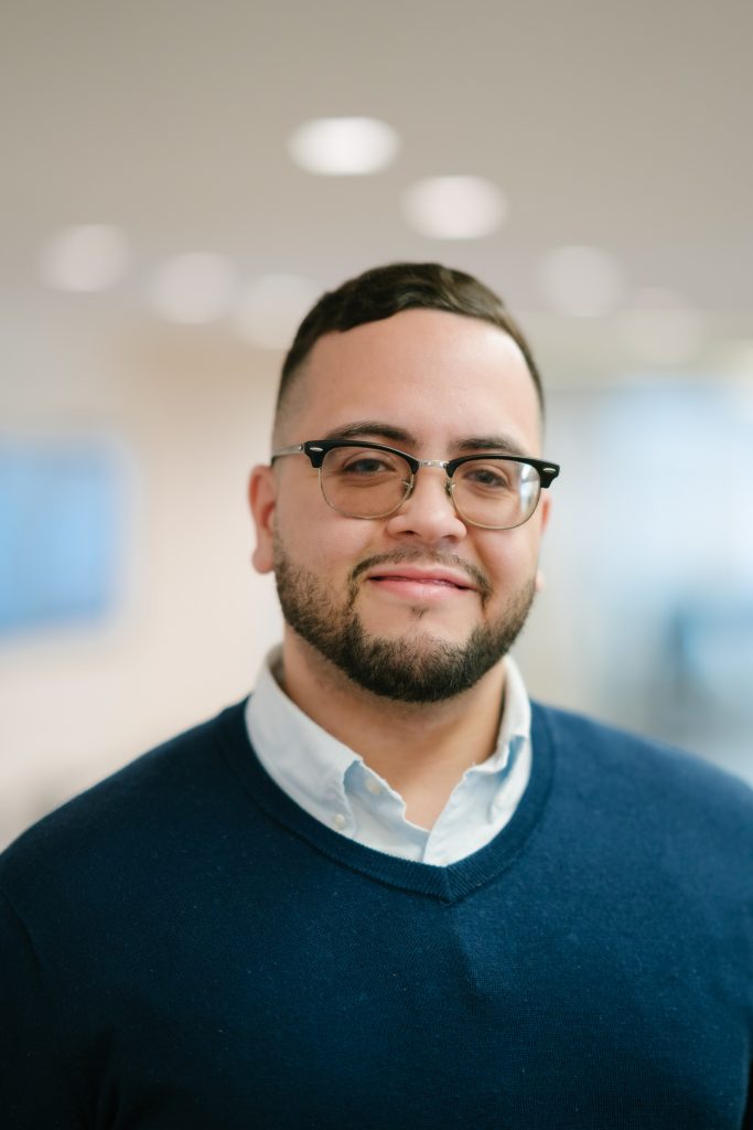 John Nunez headshot. He is wearing a Navy sweater, with button up dress shirt underneath. He is also wearing glasses.