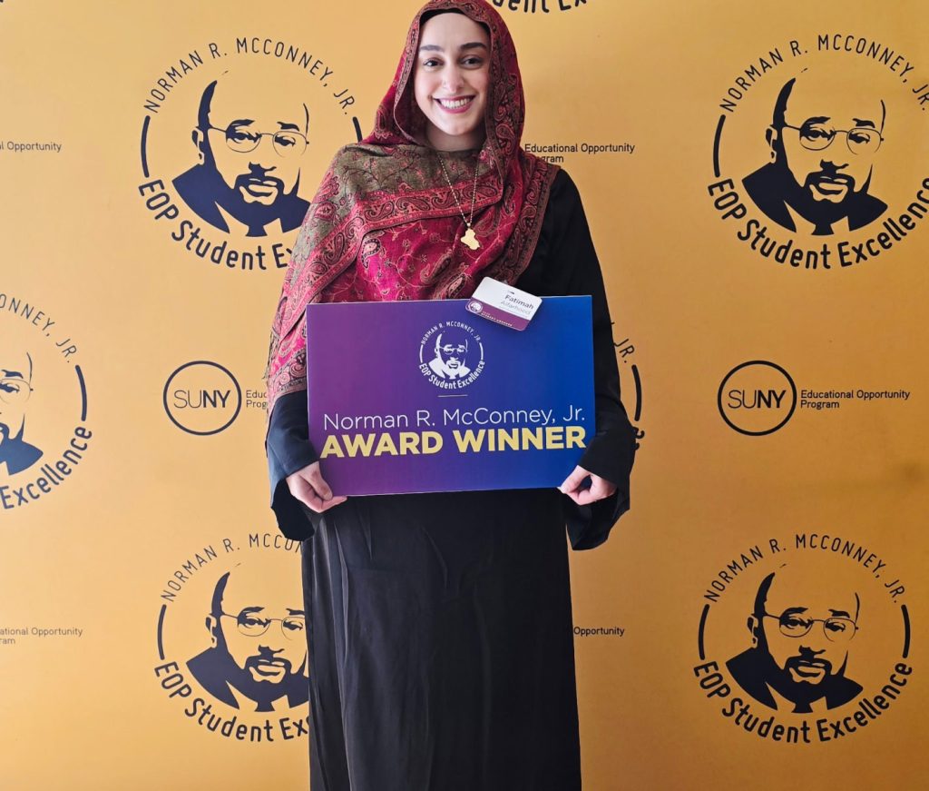 Fatimah Alfarhood wearing a black dress and red patterned hijab holding a sign saying "Norman R. McConney Jr. Award Winner"